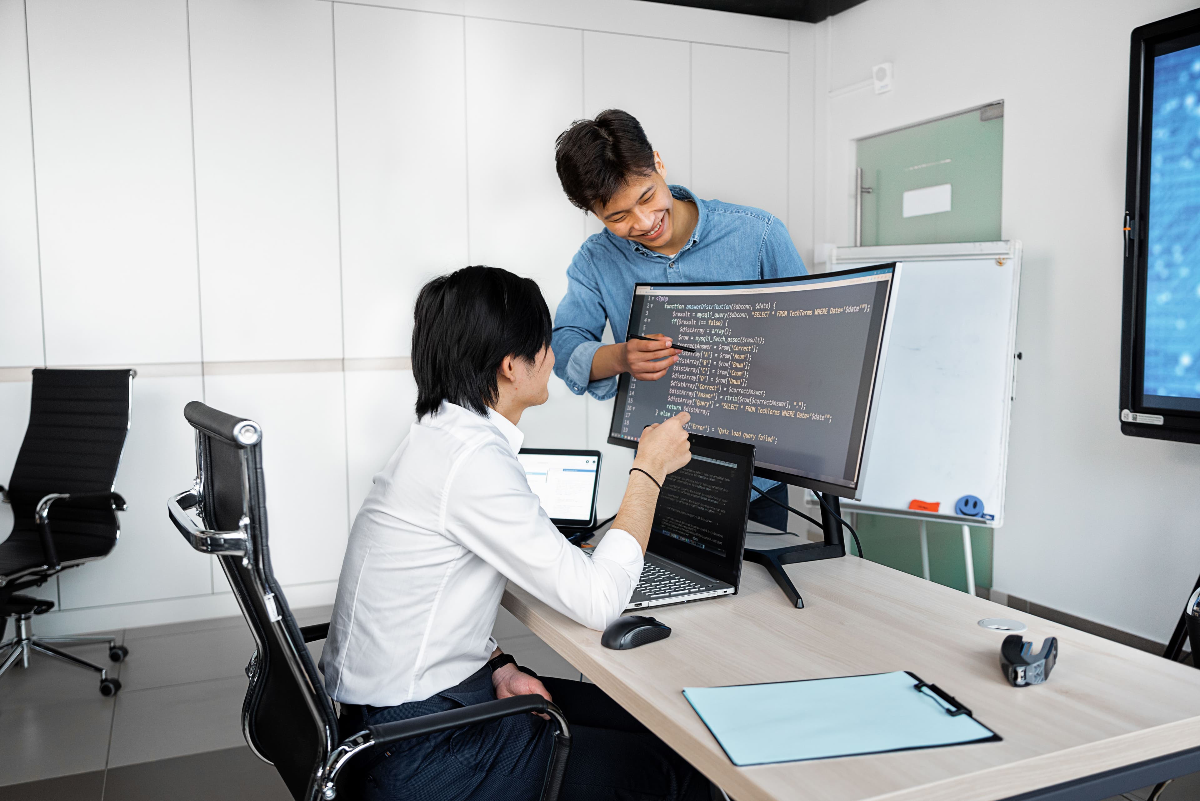 Due colleghi davanti a un monitor con del codice: uno seduto alla scrivania, l’altro che indica lo schermo