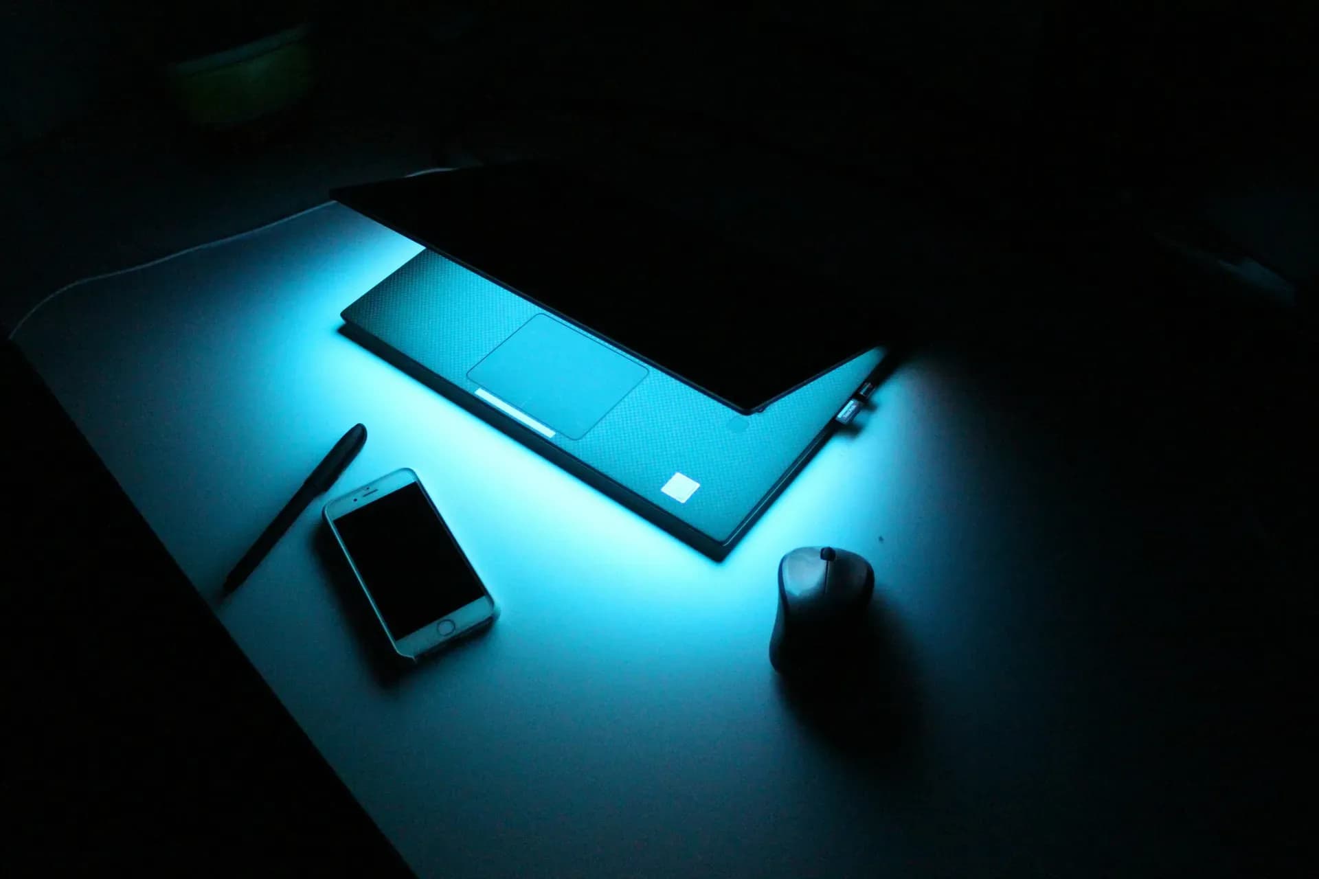 Scrivania al buio con un laptop acceso, uno smartphone e un mouse